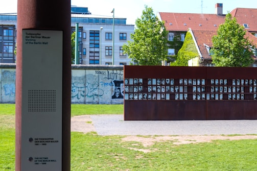 A memorial installation with a metal structure displaying numerous black-and-white portraits, set against the backdrop of a building with a red-tiled roof and a section of the graffiti-covered Berlin Wall. A plaque with inscriptions is also visible in the foreground, providing information about the victims of the Berlin Wall.