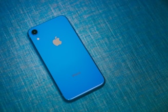 A blue smartphone with a prominent Apple logo on its back lies on a textured, blue surface. The phone's camera and flashlight are visible in the top left corner.