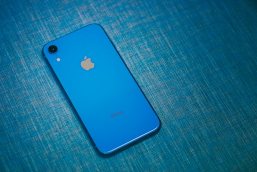 A blue smartphone with a prominent Apple logo on its back lies on a textured, blue surface. The phone's camera and flashlight are visible in the top left corner.