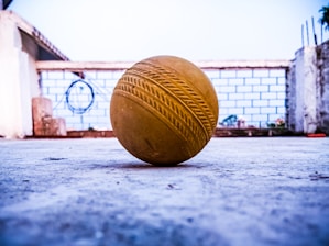 A yellow cricket ball is placed on a concrete surface, with a blurred background that includes a white brick wall and a few scattered objects.