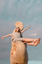 smiling woman raising both hands sitting on statue under blue and white skies