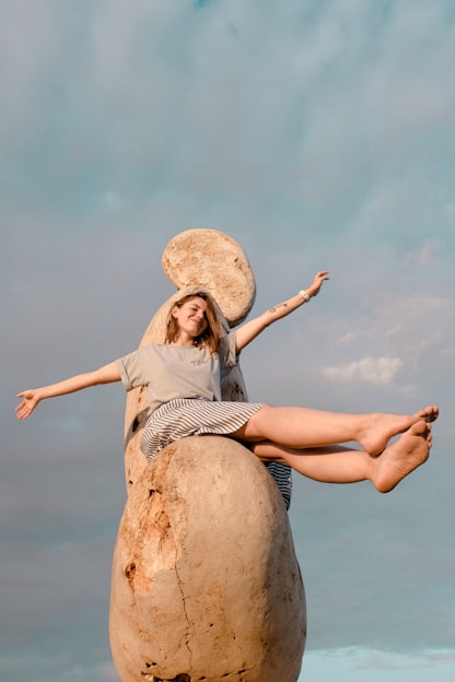smiling woman raising both hands sitting on statue under blue and white skies