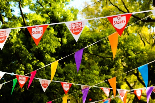 Colorful triangular flags with various colors like red, orange, purple, and blue are strung across in front of lush green trees. Some flags bear the logo 'Sagres.'