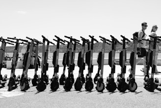 A row of electric scooters is neatly lined up on a paved surface, with a backdrop of a sunlit promenade. Two people walk by in the background, enjoying the open and relaxed atmosphere of what appears to be a coastal or lakeside setting. The landscape includes distant hills under a bright, clear sky.