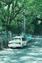 A white taxi is parked on a quiet, winding road surrounded by lush green trees. There is a fence and a prohibition road sign nearby. The scene is urban yet feels tranquil due to the dense greenery.