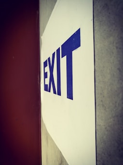 A sign with the word 'EXIT' in blue capital letters is attached to a wall. The sign is oriented vertically, and the background consists of a white surface and a brown-toned wall.
