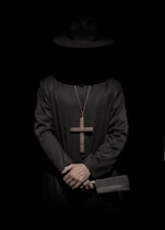 A person dressed in a black robe and hat, wearing a large cross necklace. The hands are clasped together, holding a cleaver with a prominent blade. The overall lighting is dark and shadowy, creating a mysterious and ominous atmosphere.