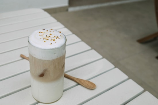A glass of creamy coffee topped with frothy milk and small brown sprinkles, placed on a light-colored slatted table. A wooden spoon lies next to the glass, and the background is softly blurred.