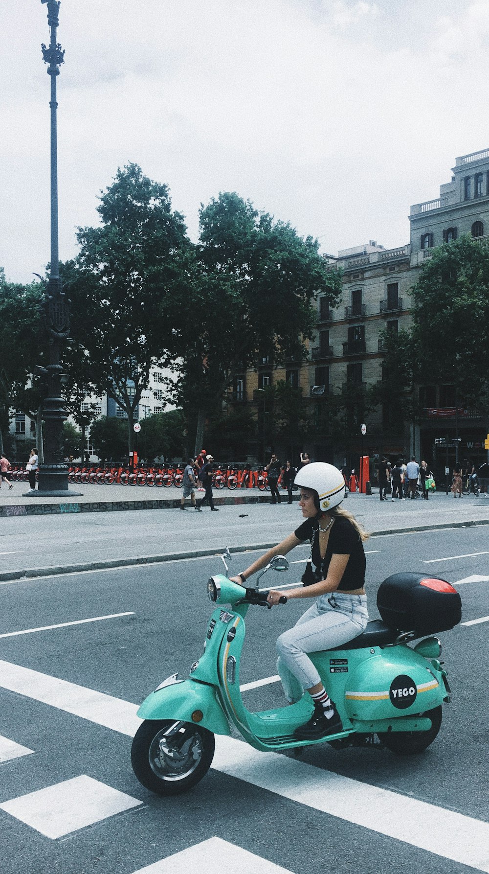 Mujer montando scooter verde cerca de la carretera que ve a la gente y los edificios bajo cielos blancos