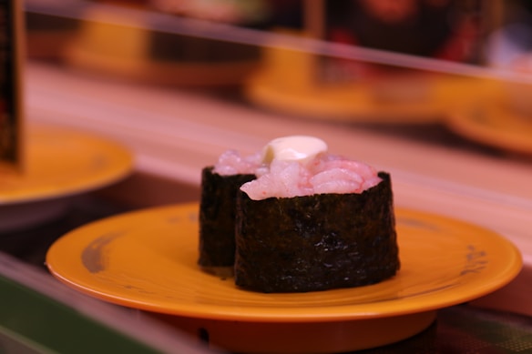 Two pieces of sushi with seaweed wrapping, topped with what appears to be cream or mayo, are placed on a rotating conveyor belt. The sushi sits on a bright yellow plate. The background is blurred, indicating a setting likely within a sushi restaurant with similar plates visible at different distances.