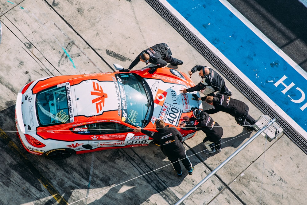 Cinq hommes debout et poussant une voiture de course blanche et orange sur la route