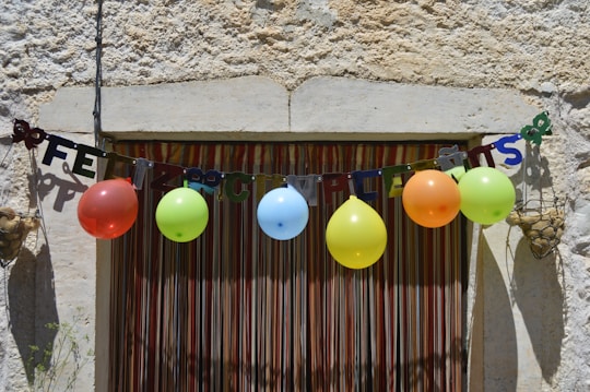 A rustic wall with a colorful 'Feliz Cumplea&ntilde;os' banner strung across, complemented by a row of vibrant balloons in various colors. The setting suggests a festive, celebratory atmosphere.