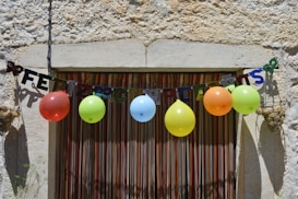 A rustic wall with a colorful 'Feliz Cumplea&ntilde;os' banner strung across, complemented by a row of vibrant balloons in various colors. The setting suggests a festive, celebratory atmosphere.