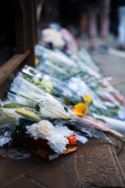 A collection of flower bouquets placed on the ground, possibly as part of a memorial or tribute. The flowers include white lilies, white chrysanthemums, and yellow gerberas, wrapped in clear plastic. A small orange-red box is also visible among the flowers, resting on a stone-paved ground.