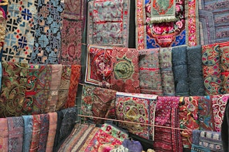 A vibrant array of richly embroidered and intricately patterned textiles are displayed, covering a wall with a variety of ornate designs. The fabrics feature a mix of geometric and floral motifs, with colors ranging from deep blues and reds to bright pinks and greens. Each textile appears to be hand-crafted with detailed stitching and unique patterns.