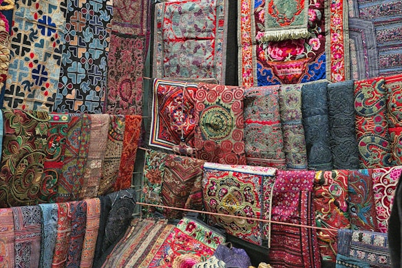 A vibrant array of richly embroidered and intricately patterned textiles are displayed, covering a wall with a variety of ornate designs. The fabrics feature a mix of geometric and floral motifs, with colors ranging from deep blues and reds to bright pinks and greens. Each textile appears to be hand-crafted with detailed stitching and unique patterns.