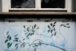 A wall mural depicting branches with green leaves and delicate lines extending across a panel. Above the mural, a window reflects a cloudy sky. Two drawn eyes are positioned above the branches, adding a surreal touch.
