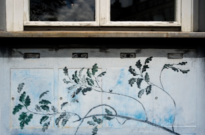 A wall mural depicting branches with green leaves and delicate lines extending across a panel. Above the mural, a window reflects a cloudy sky. Two drawn eyes are positioned above the branches, adding a surreal touch.