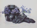 A motorcycle helmet filled with vibrant purple flowers sits beside a pair of black gloves. In the background, a container filled with the same type of flowers adds a decorative touch. The flowers contrast beautifully against the dark helmet and gloves, lending a soft yet striking appearance to the scene.