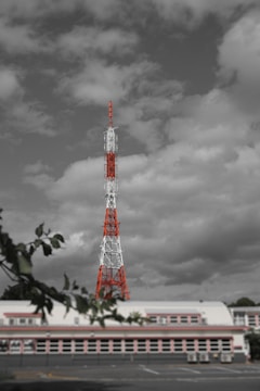 A tall radio or television transmission tower with red and white sections stands prominently against a backdrop of cloudy skies. The structure dominates the scene and is surrounded by a partially visible building and foreground tree branches.
