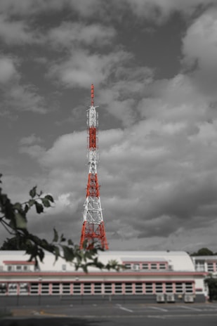 A tall radio or television transmission tower with red and white sections stands prominently against a backdrop of cloudy skies. The structure dominates the scene and is surrounded by a partially visible building and foreground tree branches.