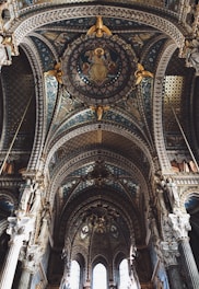 Elaborate and ornate cathedral ceiling featuring intricate designs, religious iconography, and a rich interplay of colors and textures. Statues of religious figures adorn the upper columns, and the central medallion depicts a seated figure. The architecture highlights gothic and renaissance influences with detailed arches and pillar sculptures.