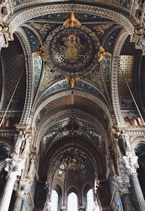 Elaborate and ornate cathedral ceiling featuring intricate designs, religious iconography, and a rich interplay of colors and textures. Statues of religious figures adorn the upper columns, and the central medallion depicts a seated figure. The architecture highlights gothic and renaissance influences with detailed arches and pillar sculptures.