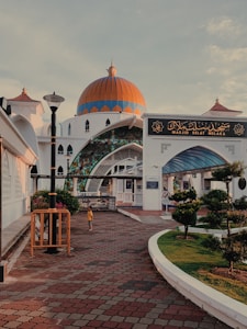 A mosque with a striking orange and blue dome stands against a partly cloudy sky. The entrance features intricate stained glass and Islamic architectural elements. A large sign with Arabic script is displayed above the entrance. The surrounding area includes tiled pathways, well-maintained hedges, and a lone figure wearing a yellow shirt walking nearby.