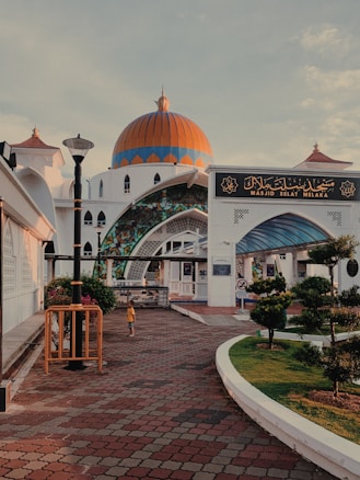 A mosque with a striking orange and blue dome stands against a partly cloudy sky. The entrance features intricate stained glass and Islamic architectural elements. A large sign with Arabic script is displayed above the entrance. The surrounding area includes tiled pathways, well-maintained hedges, and a lone figure wearing a yellow shirt walking nearby.