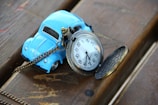 A vintage pocket watch with an ornate chain rests against a small blue toy car on a wooden surface. The watch is open, displaying its white face and black numerals.