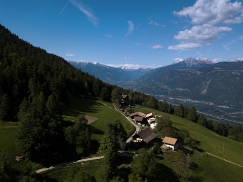 A scenic landscape with a cluster of traditional houses surrounded by lush green fields and dense forests. Snow-capped mountains are visible in the background under a partly cloudy blue sky. A narrow path winds through the greenery leading to the houses.