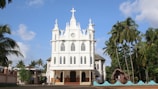 A white, ornate church structure with Gothic architectural elements, featuring pointed arches and intricate details. The church is flanked by tall palm trees under a clear blue sky. Several people are visible near the entrance, with a stone grotto on the right side.
