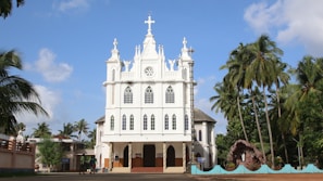 A white, ornate church structure with Gothic architectural elements, featuring pointed arches and intricate details. The church is flanked by tall palm trees under a clear blue sky. Several people are visible near the entrance, with a stone grotto on the right side.