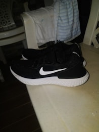 A pair of black athletic shoes with a white swoosh logo, resting on a light-colored table. In the background, a chair draped with a folded blanket featuring light blue and white stripes is visible.