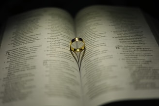 A gold ring rests on an open book, with its shadow forming a heart shape on the pages. The text on the book is in a language with a distinct script, possibly Malayalam.