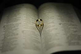 A gold ring rests on an open book, with its shadow forming a heart shape on the pages. The text on the book is in a language with a distinct script, possibly Malayalam.