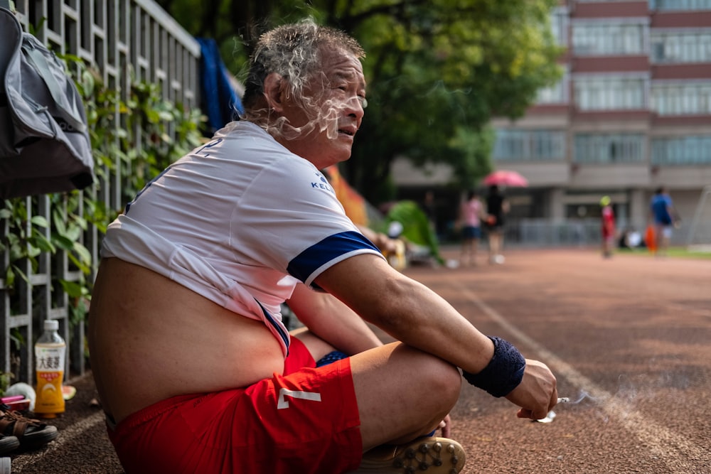 man in white t-shirt and red shorts sitting on ground smoking cigarette