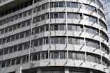 A modern multi-story building with a curved facade featuring rows of rectangular windows. The structure has an industrial appearance with vertical and horizontal lines created by metallic beams and panels.