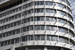 A modern multi-story building with a curved facade featuring rows of rectangular windows. The structure has an industrial appearance with vertical and horizontal lines created by metallic beams and panels.