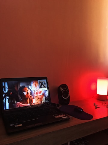 A laptop displaying a woman wearing sunglasses with a neon sign reflection on the screen sits on a wooden desk. Beside the laptop is a black speaker and a dark mouse pad. A small lamp emits a warm red light on the right-hand side of the desk, with earbuds lying close by.