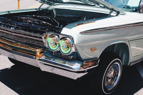 A vintage car is displayed with its hood open, revealing parts of the engine. The car features distinctive round headlights with green-tinted covers and a well-maintained chrome trim. The bodywork is a pale pastel color with a classic design emblem on the fender.