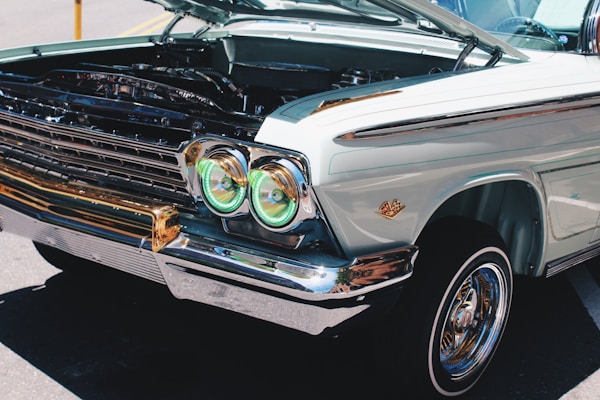 A vintage car is displayed with its hood open, revealing parts of the engine. The car features distinctive round headlights with green-tinted covers and a well-maintained chrome trim. The bodywork is a pale pastel color with a classic design emblem on the fender.