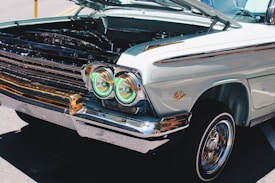 A vintage car is displayed with its hood open, revealing parts of the engine. The car features distinctive round headlights with green-tinted covers and a well-maintained chrome trim. The bodywork is a pale pastel color with a classic design emblem on the fender.