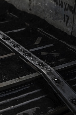 Rusty metal rails stretch over a dark, gravelly surface with water droplets adding a sense of dampness. The background features a concrete wall with faded graffiti, creating an urban, abandoned atmosphere.