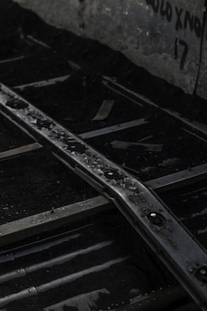 Rusty metal rails stretch over a dark, gravelly surface with water droplets adding a sense of dampness. The background features a concrete wall with faded graffiti, creating an urban, abandoned atmosphere.