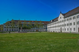 A large white building with green shutters and a brown roof is bordered by well-maintained grass and a clear blue sky. Trees line the front of the building, which has a modern glass extension connecting two wings.
