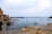 A serene coastal scene with a historic stone structure featuring a clock tower to the left and numerous sailboats and yachts anchored in calm waters. The sky is overcast, adding a tranquil atmosphere, while seagulls fly above. People are seen walking along the path near the water's edge.