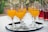 Several glasses filled with orange juice are arranged on a black tray covered with a white paper napkin. The foreground features this tray placed on a table with a subtle patterned tablecloth. In the background, a few glasses containing a red beverage are visible, adding contrast against the greenery.