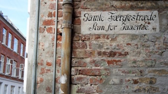 A weathered brick wall with an old sign in ornate text hangs above, displaying the words 'Gamle F&aelig;rgestr&aelig;de kun for gaaende'. A metal drainpipe runs vertically along the wall. Adjacent to this scene, there are red brick buildings with white-framed windows.