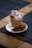 A glass of hot chocolate topped with marshmallows and garnished with a dusting of cocoa powder sits on a white saucer. Beside it lies a chocolate bar partially cut open to reveal a caramel and nougat filling. The setup rests on a wooden table with a dark textured placemat.